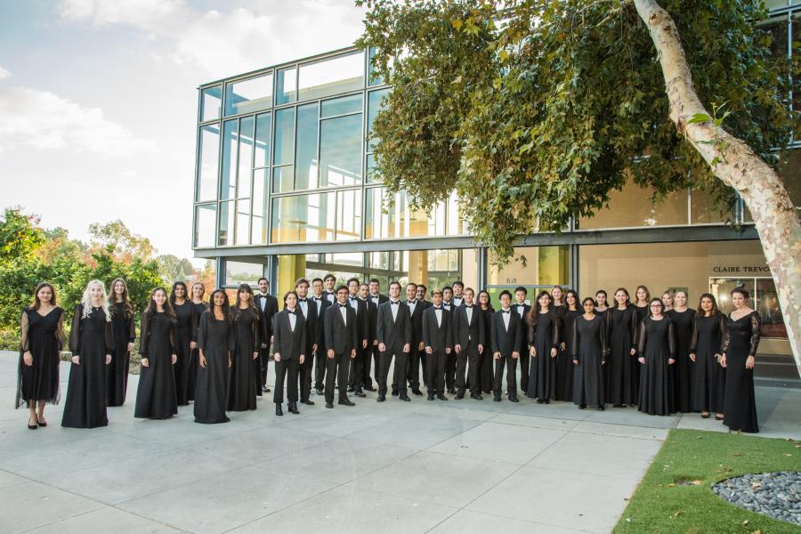 Outdoor group photo of a concert choir in black formal attire lined up on a walkway in front of a modern glass building, framed by a large tree and afternoon light.
