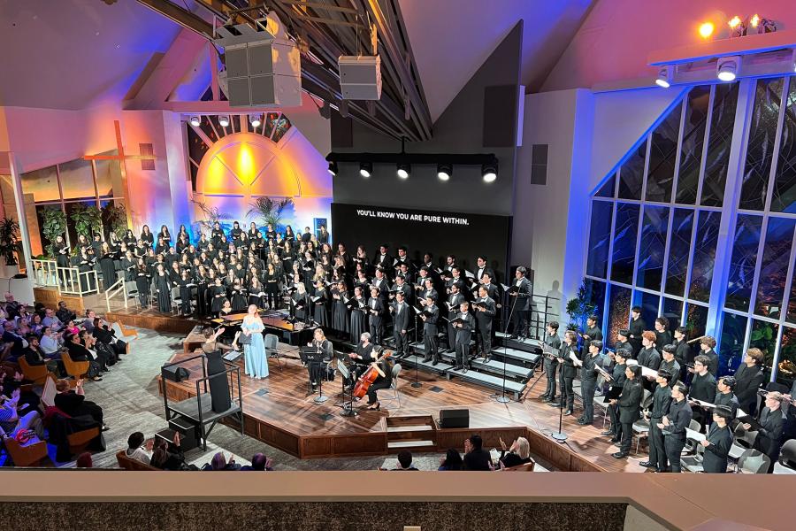 High-angle view of a choir concert in a bright, church-like venue with blue and amber lighting; singers on risers flank the stage while an audience watches below.