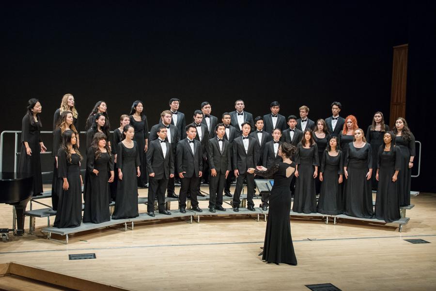 Concert choir in black dresses and tuxedos stands on tiered risers while a conductor in a long black dress leads from the floor; grand piano at left, dark backdrop.