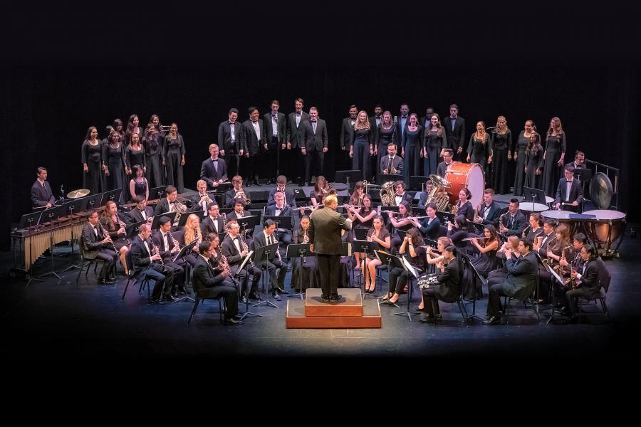 Wind ensemble and choir on a dark stage with a conductor on a center podium; musicians seated with music stands, choir on risers behind, and percussion instruments at rear.