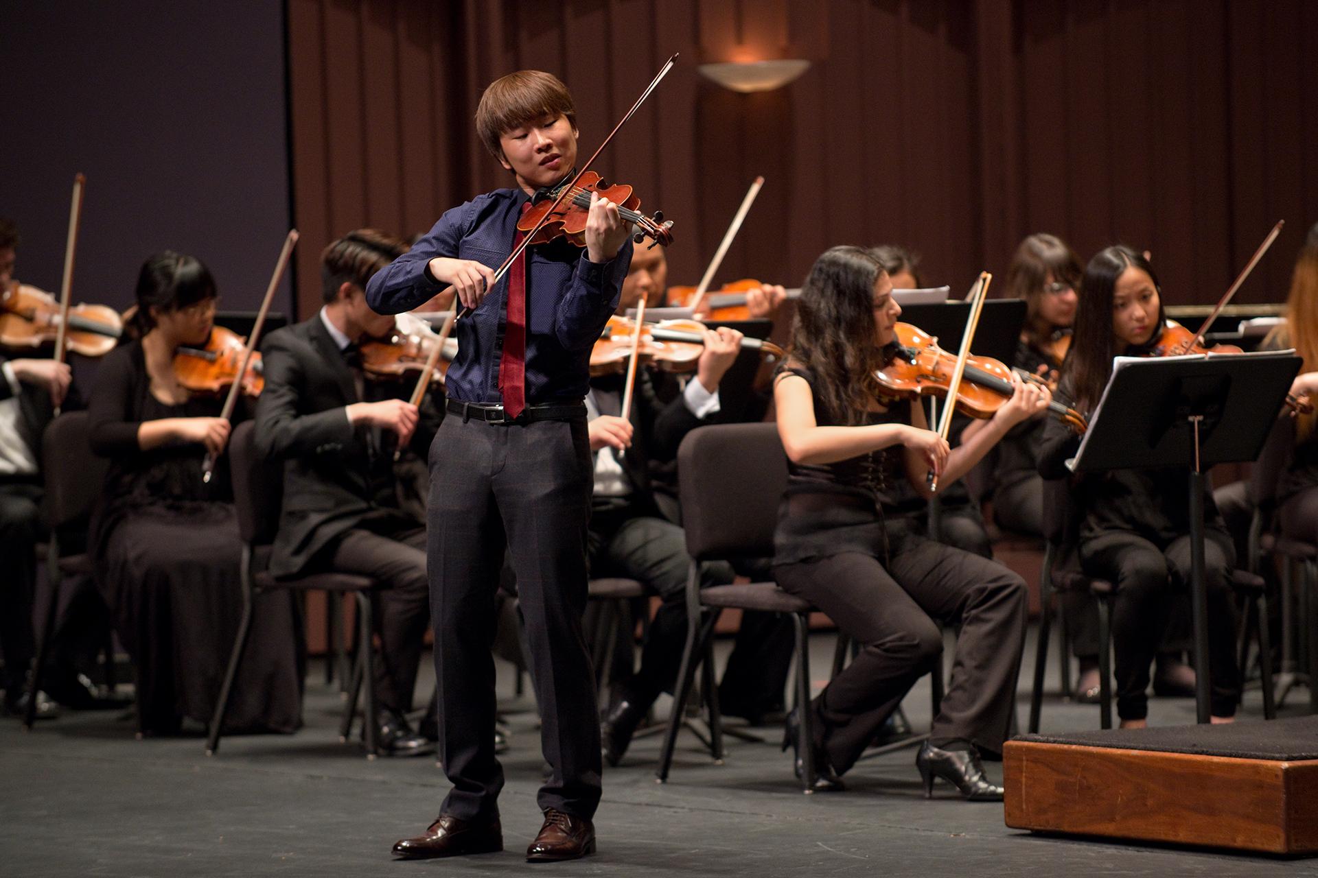 Alt text: A violin soloist stands at the front of a concert stage, playing with eyes partly closed, while seated orchestra musicians perform behind them with music stands and violins under warm stage lighting.