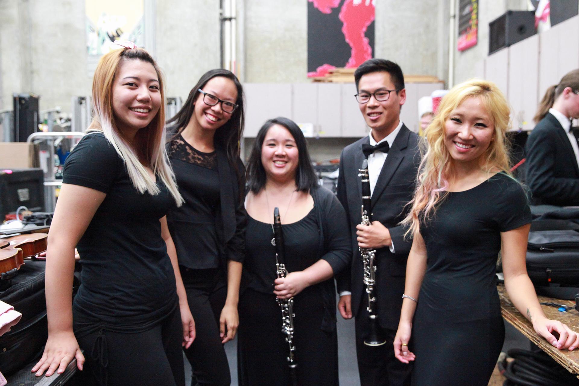 Five smiling musicians pose backstage in a rehearsal/production area; two hold clarinets, one person wears a tuxedo, and instrument cases and equipment are visible around them.