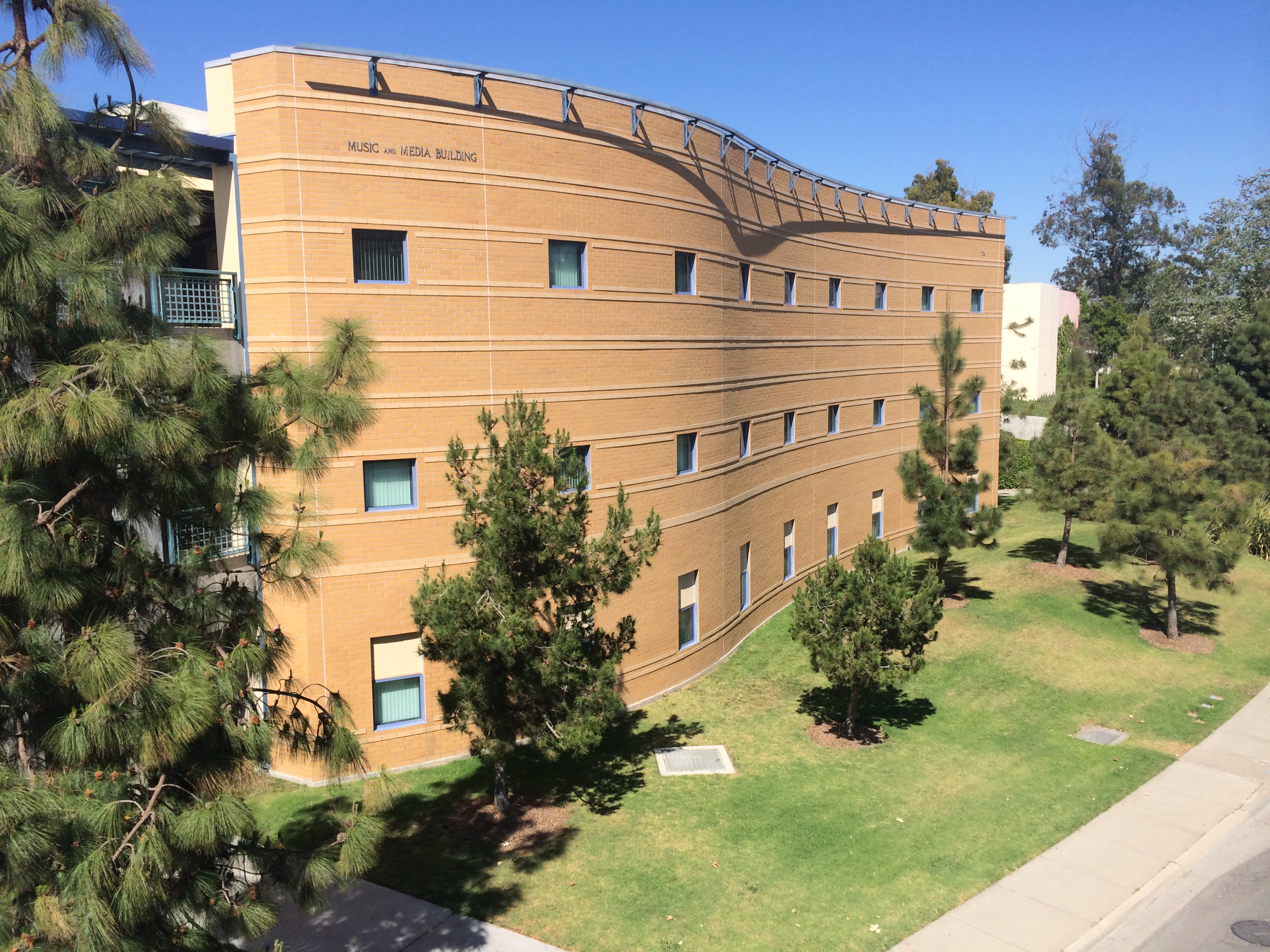 Exterior view of the Music and Media Building, a curved brick academic structure surrounded by pine trees and lawn under a clear blue sky.