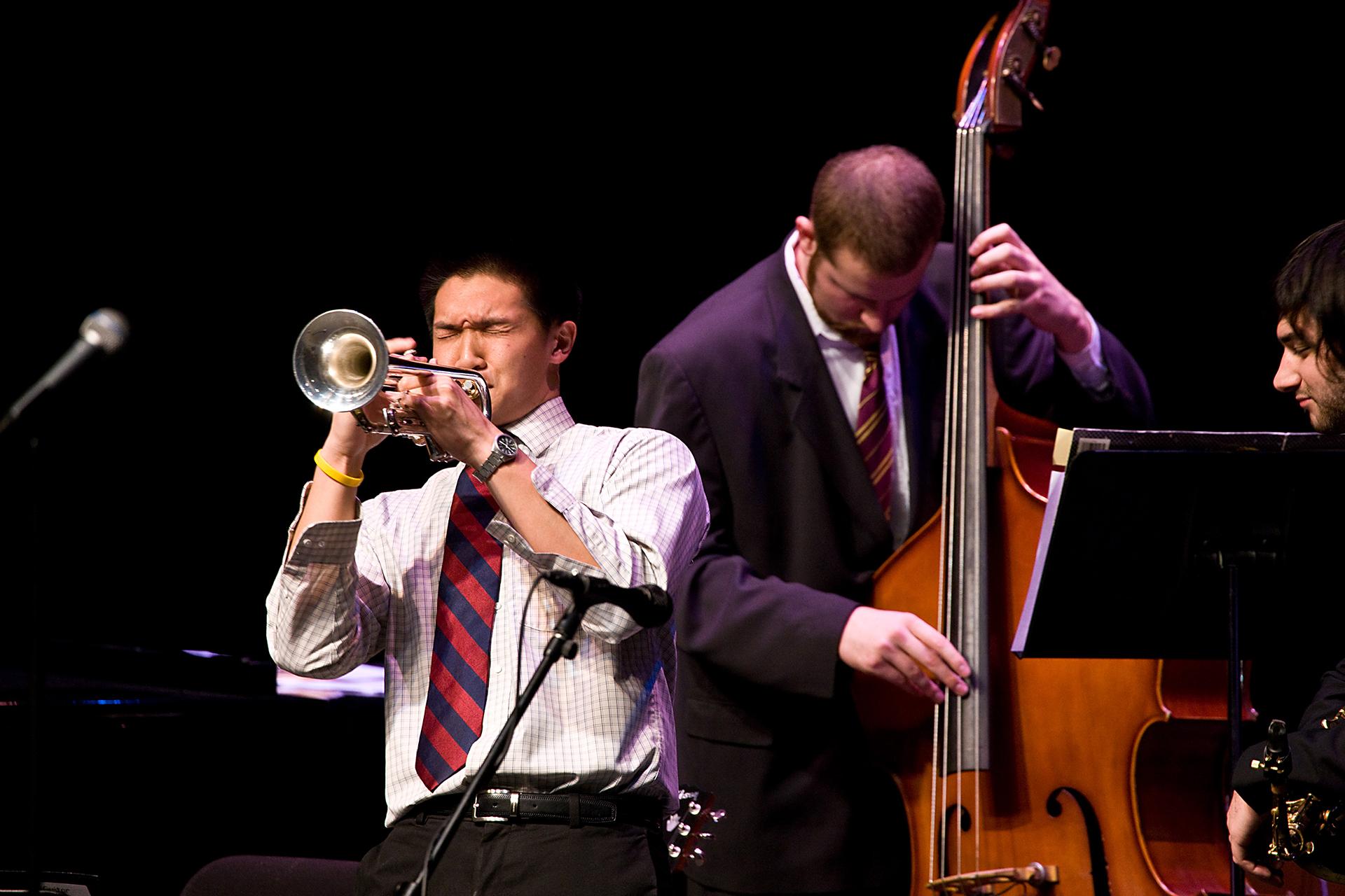 On a dark stage, a trumpeter plays with eyes closed while a bassist in a suit performs behind him next to a music stand and microphones.