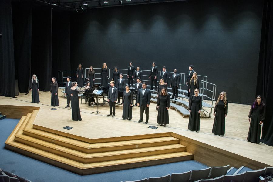 A formal choir ensemble dressed in black attire stands on tiered risers on a wooden stage with a piano. Blue auditorium seating is visible in the foreground with dramatic stage lighting above.