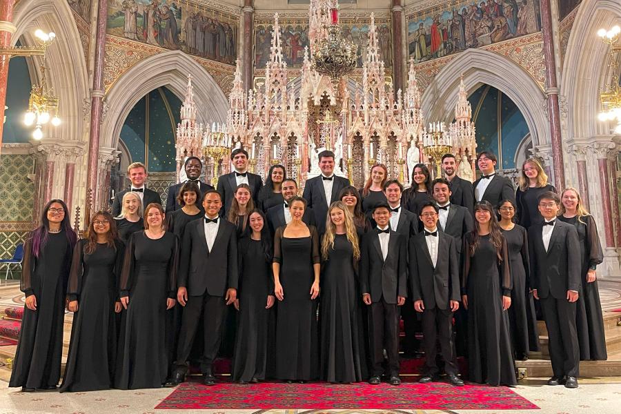 A formal choir group in black attire poses in rows inside an ornate Gothic church sanctuary, standing before an elaborate altar with tall spires, crystal chandeliers, and religious frescoes.