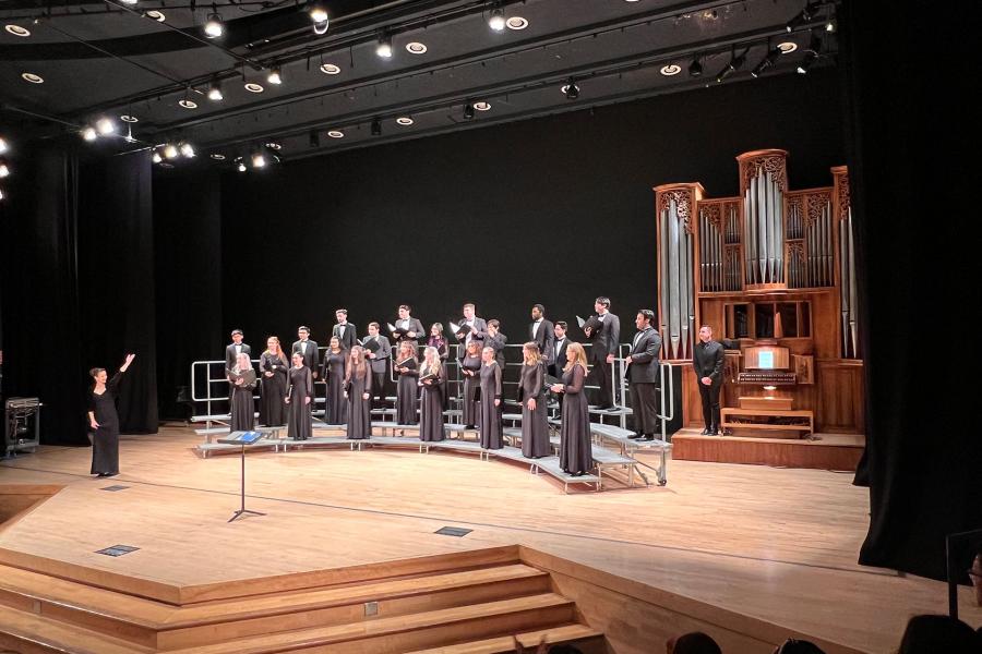 A choir ensemble in formal black attire performs on stage with tiered risers, led by a conductor on the left. A large wooden pipe organ stands on the right. The stage features professional lighting and a wooden floor in a concert hall setting.