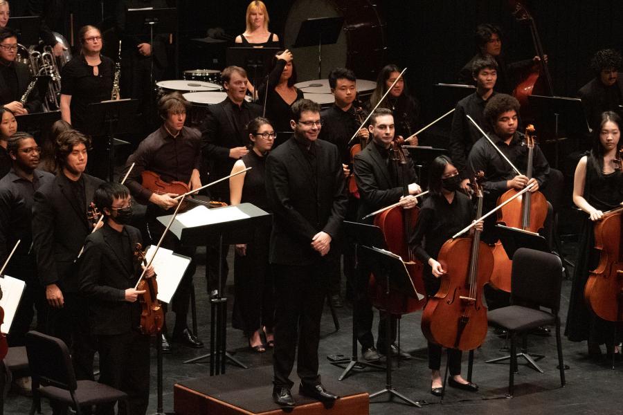 Orchestra on a dim stage during curtain call; conductor and musicians in black stand near music stands while cellists and other players hold instruments; percussion and winds visible behind.