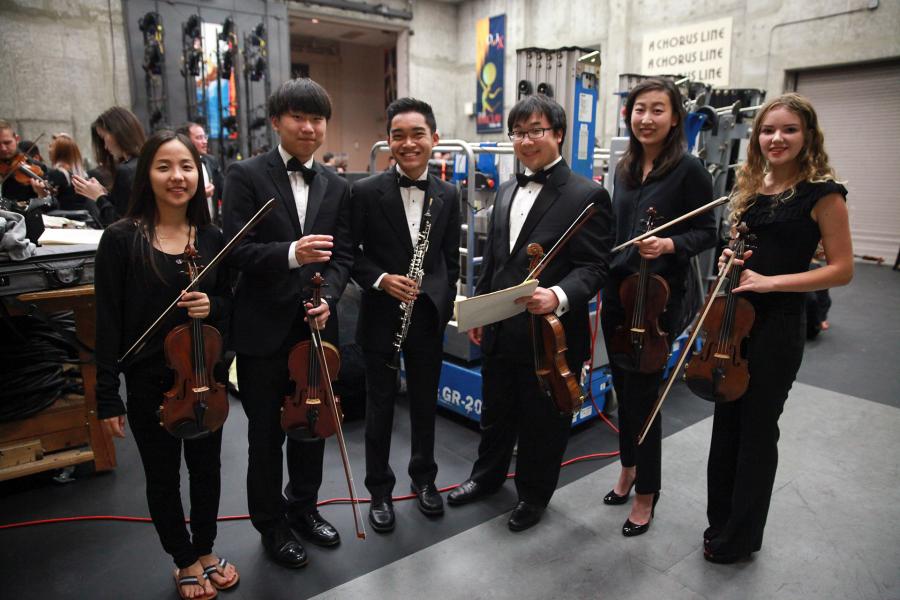 Six student musicians pose backstage in formal black; several hold violins/viola, one holds an oboe and another holds sheet music; stage rigging, cases, and equipment surround them.