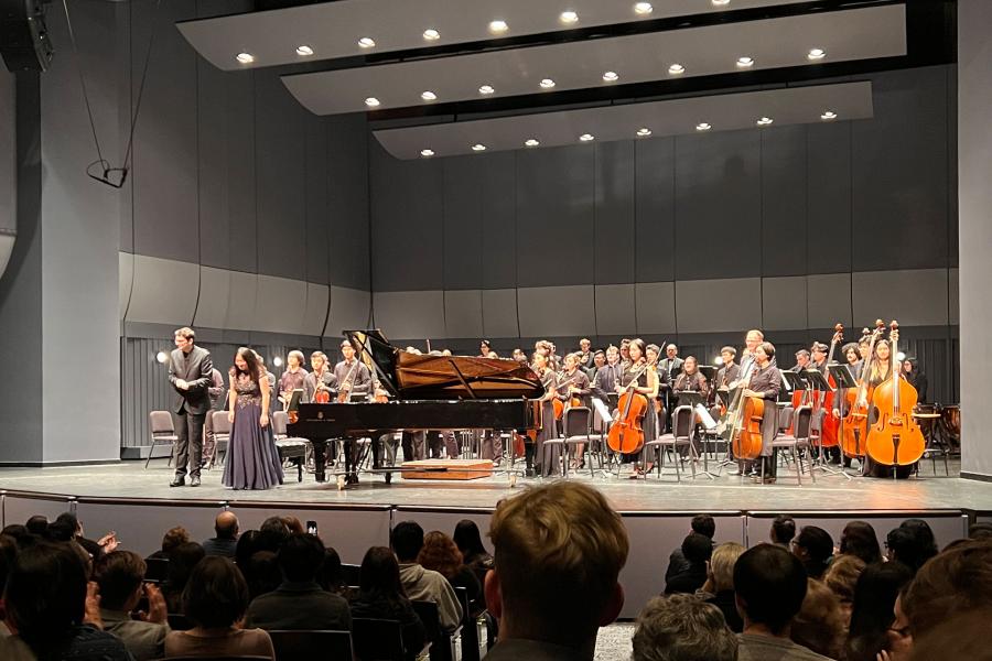 Wide audience view of a concert hall stage: grand piano at center as conductor and soloist bow; full orchestra stands behind with strings and double basses under bright ceiling lights.