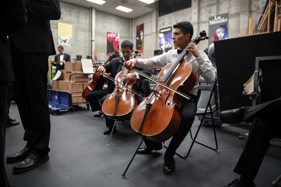 Backstage rehearsal scene with two seated cellists in formal attire; one plays while another tunes, surrounded by cables, cases, and posters; a conductor’s legs frame the left foreground.