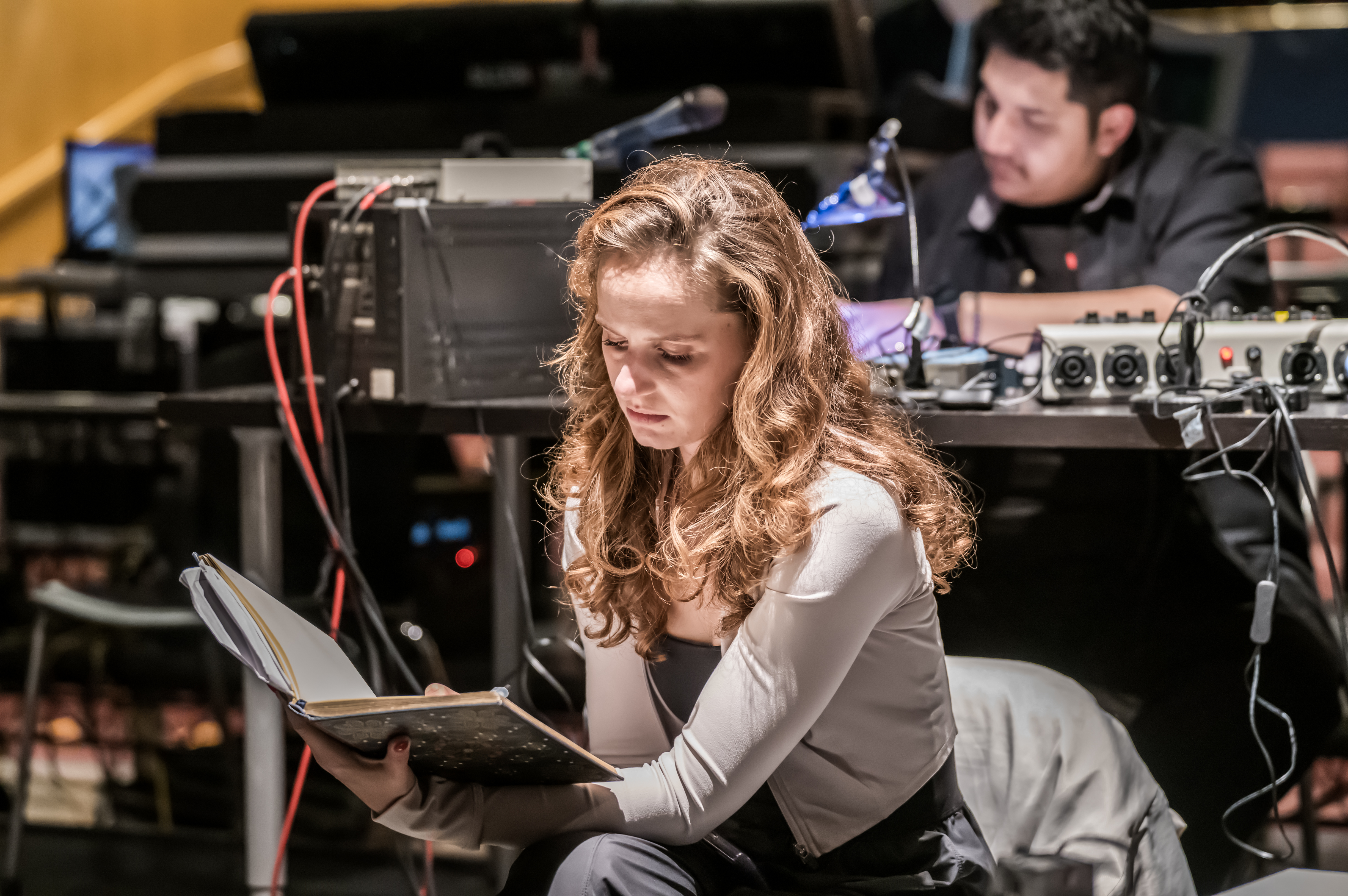 A UCI Drama student reads from a script during rehearsal while technical equipment and crew are visible in the background.