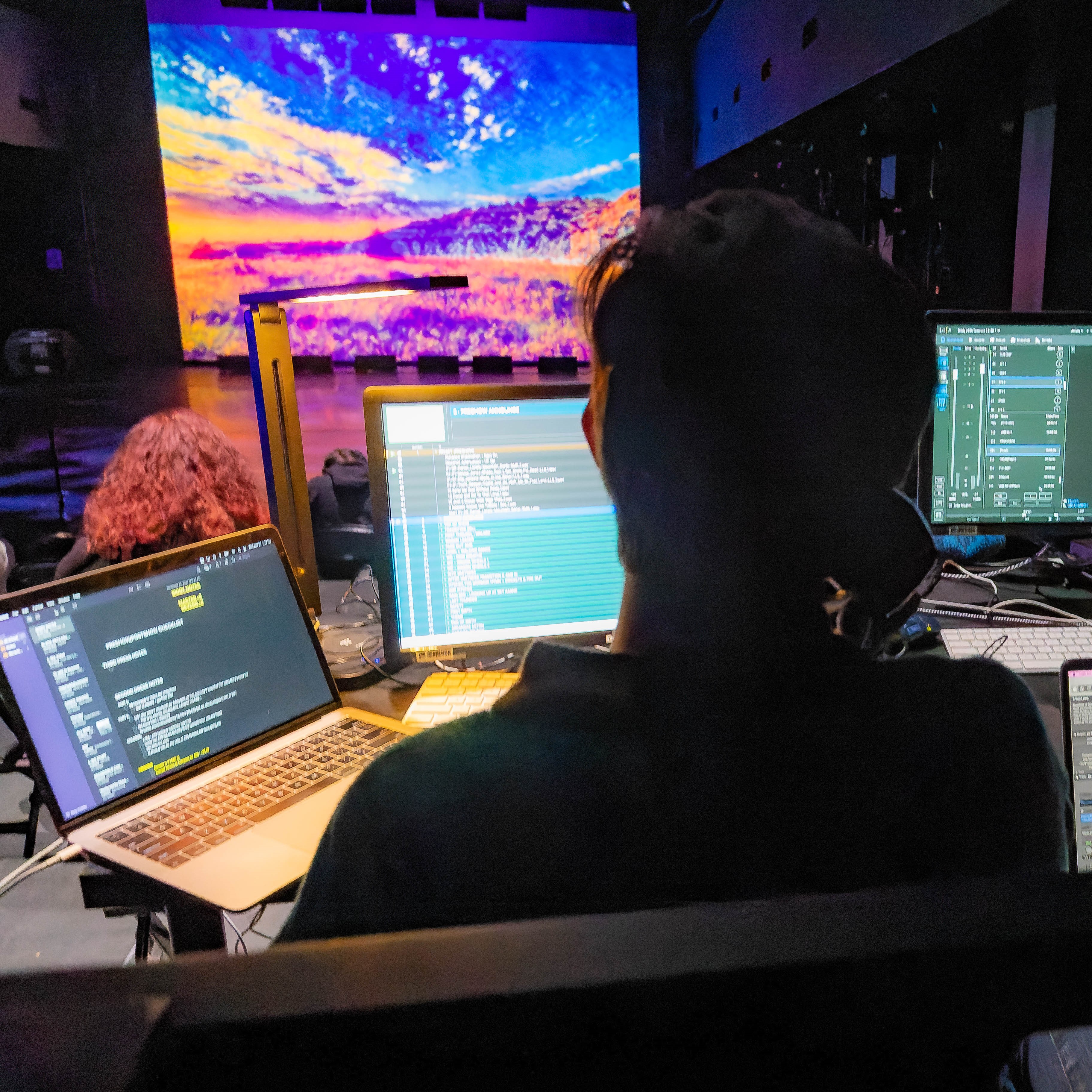 A UCI Arts student operates lighting and media controls during a production, with digital visuals projected onstage in the background.