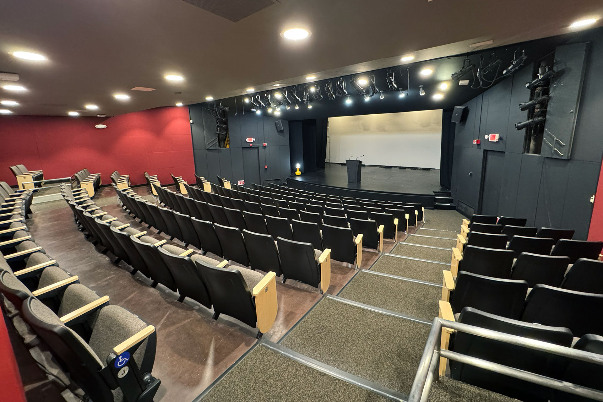 The Little Theatre: Empty proscenium-style theater with tiered seating facing a stage with a podium, overhead lighting grid and black walls with red accents