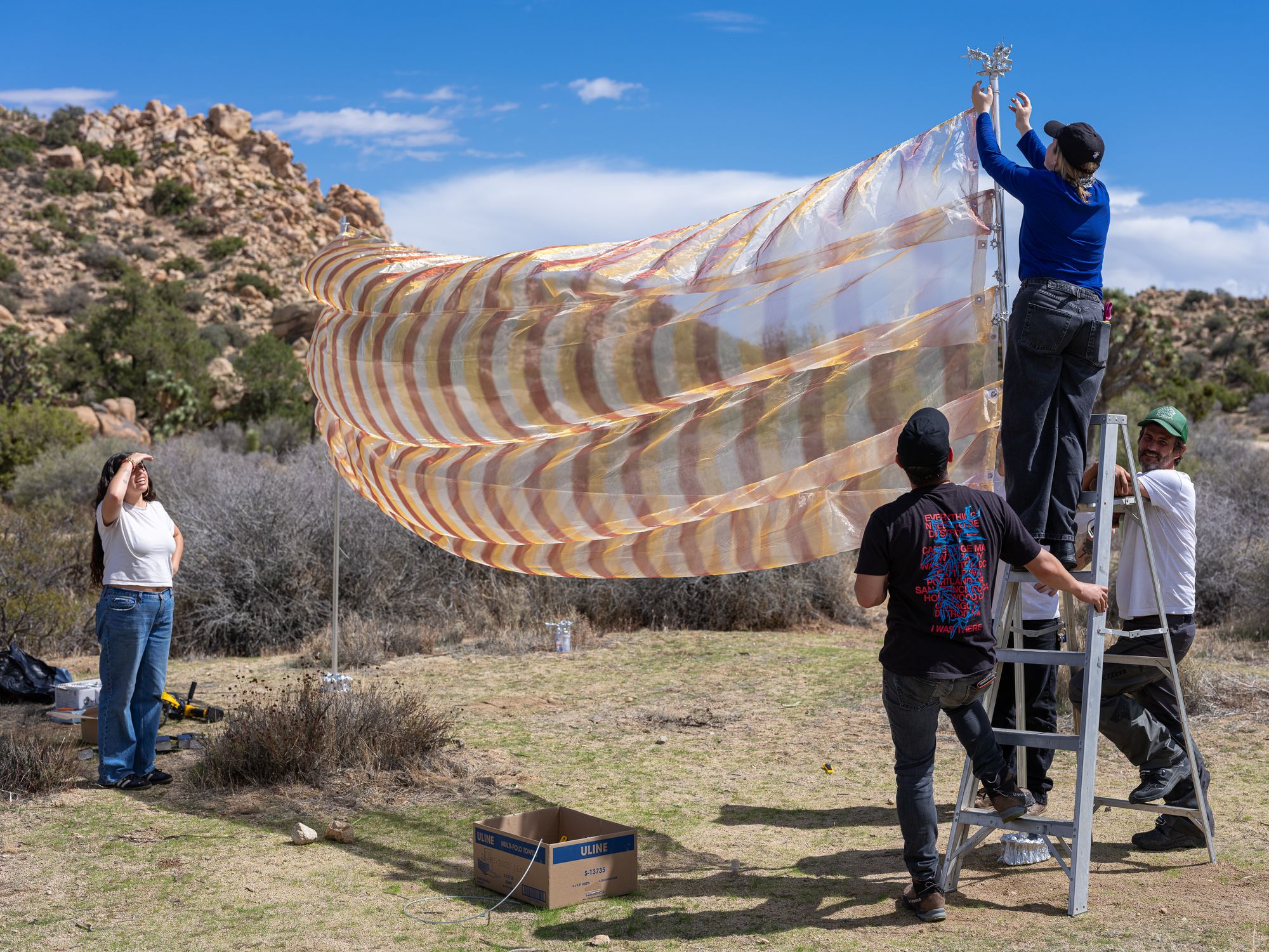 Liz Stringer and other Department of Art students at Burns Pinon participants install Stringer's work So Blue at Burns Projects, September 2025. Photo: Jesse Colin Jackson (Group works together to secure a large striped translucent fabric installation using a ladder in a rocky desert environment)