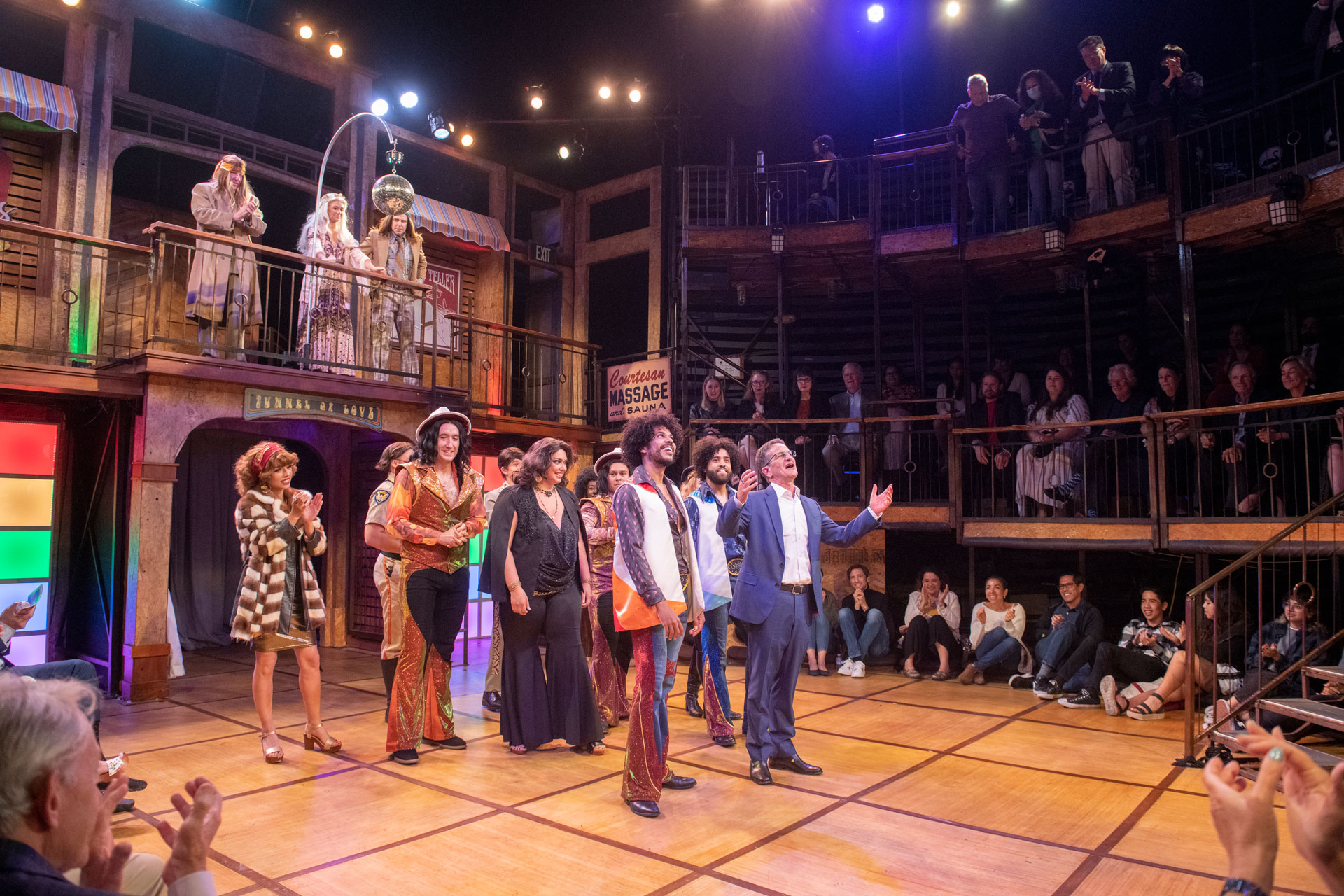 Eli Simon and cast members from Comedy or Errors stand center stage during a curtain call in a multi-level immersive theater space as audience members applaud from surrounding balconies and floor seating
