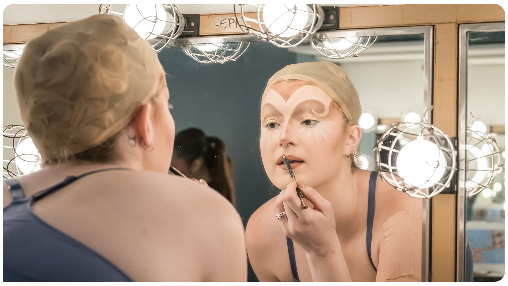Performer applies stage makeup in a dressing room mirror surrounded by lit bulbs 