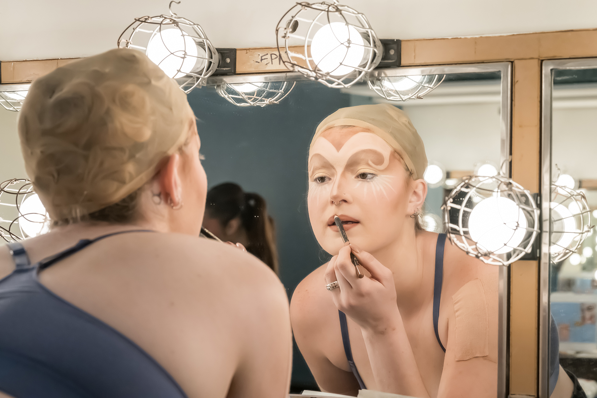 A student applies theatrical makeup in front of a lit dressing room mirror, preparing for a performance