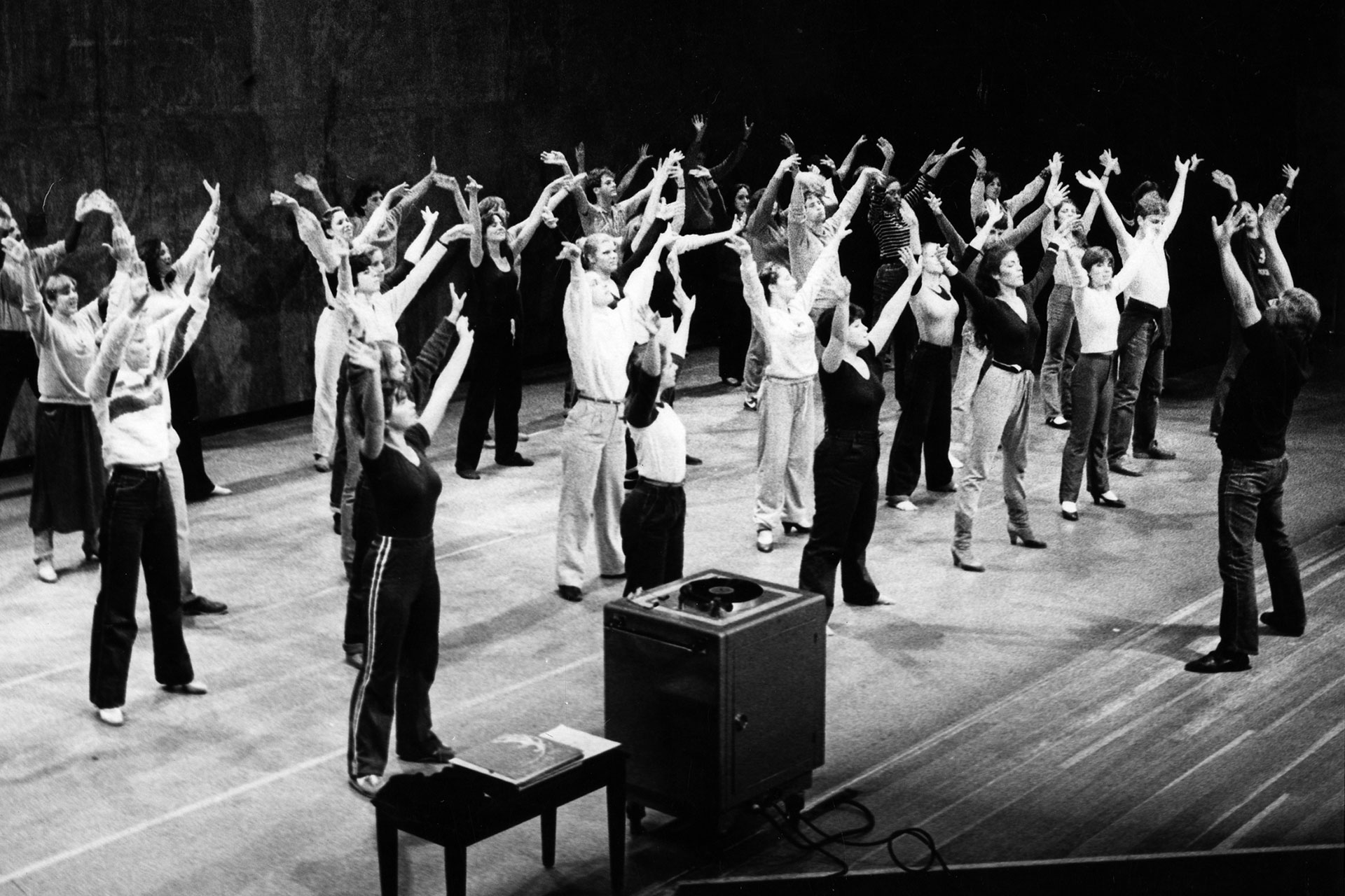 Clayton Garrison teaching class, black-and-white photo of a large group of students practicing with arms raised in a studio.