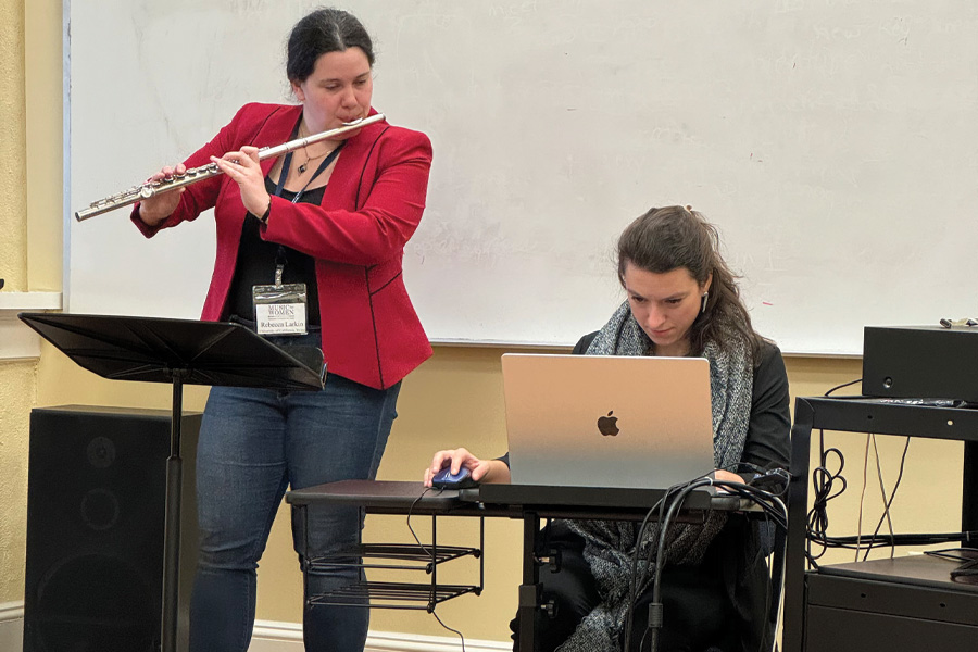Rebecca Larkin in red jacket playing flute as participant plays video game.