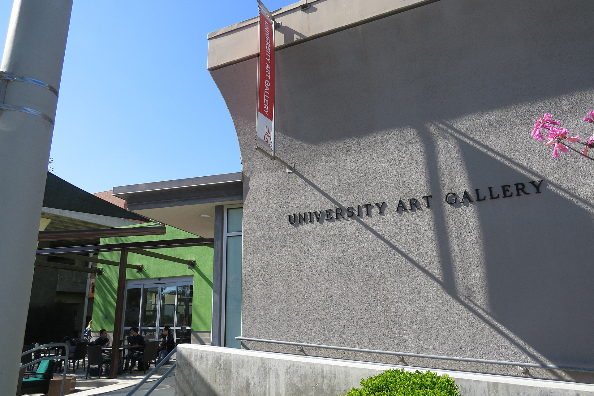 Exterior view of the University Art Gallery building with wall signage, entryway and outdoor seating area under clear daylight