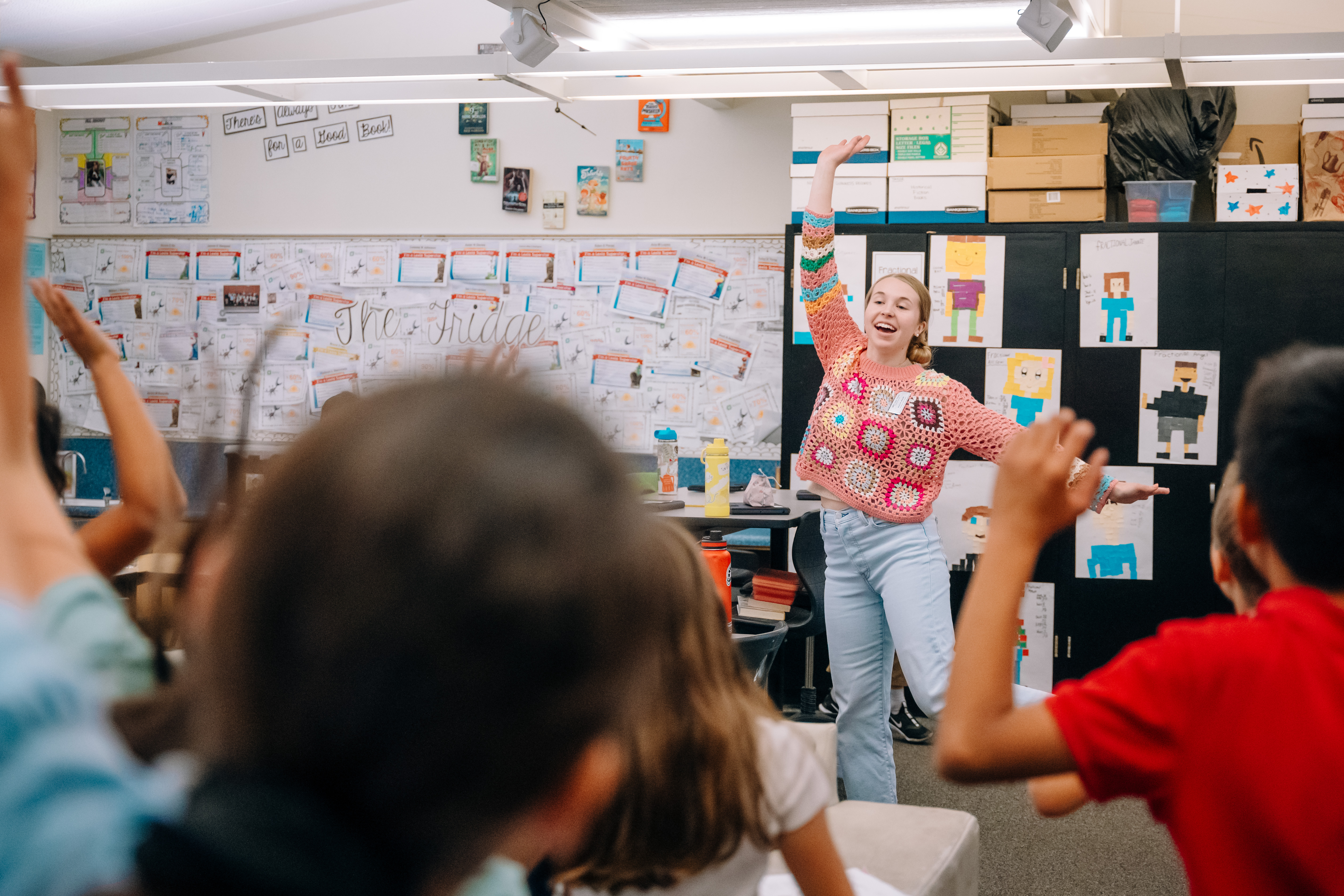 A UCI Arts student stands at the front of a classroom with her arms raised, facing elementary school students who mirror her gesture.