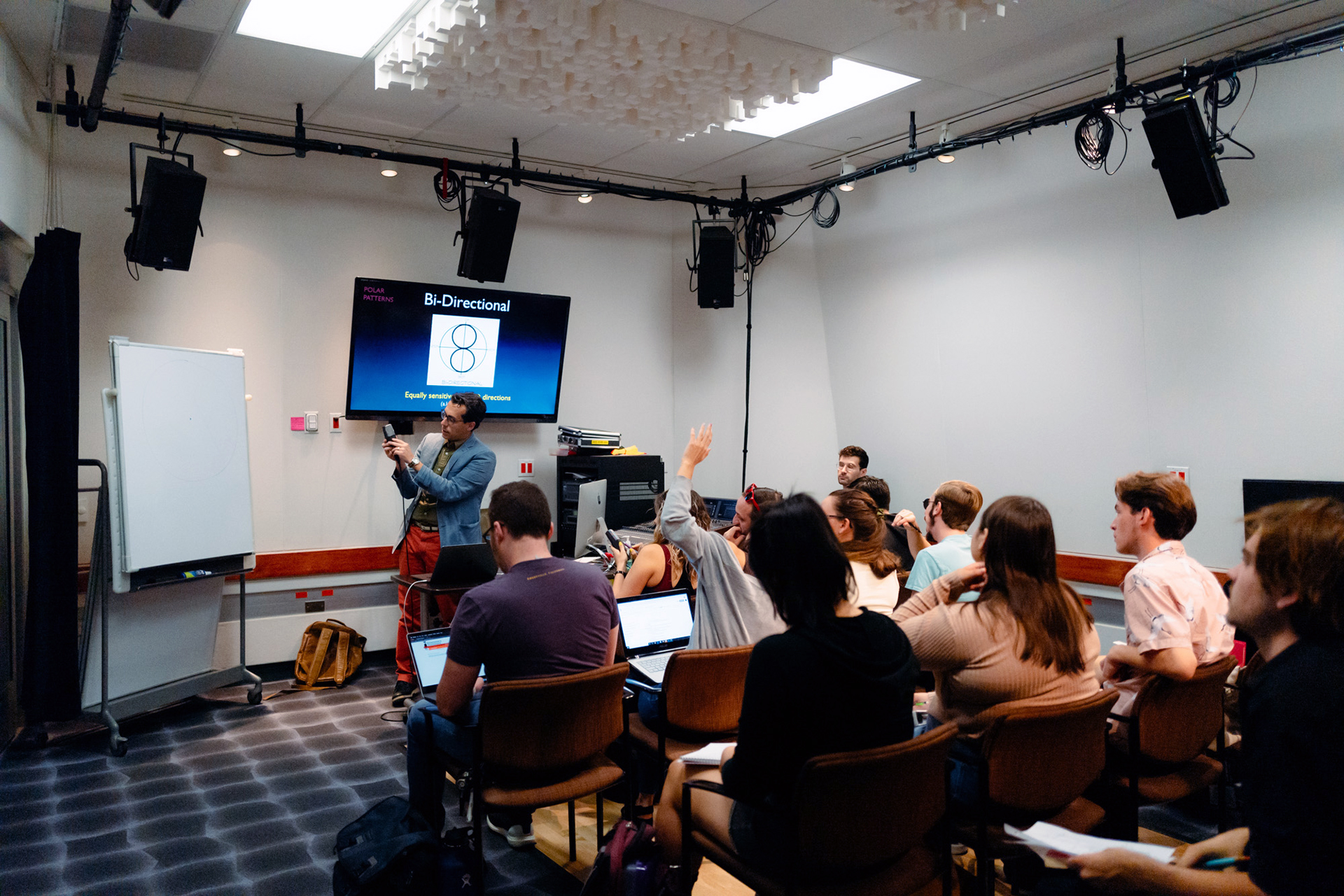 Professor Vincent Oliveri presents audio concepts using a screen display while students sit with laptops and raise hands in an interactive classroom setting