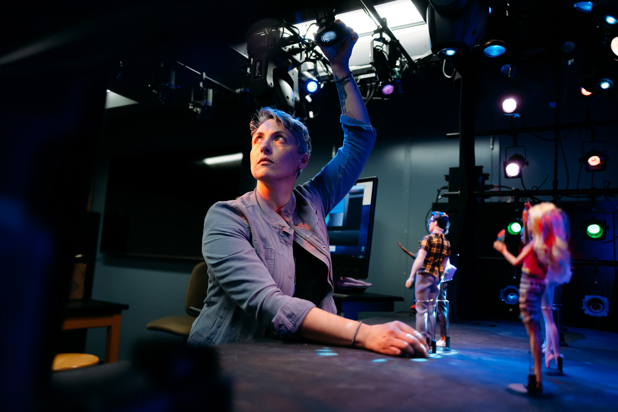 Professor Jaymi Smith adjusts overhead stage lighting in a performance lab while working with small puppet figures on a table.