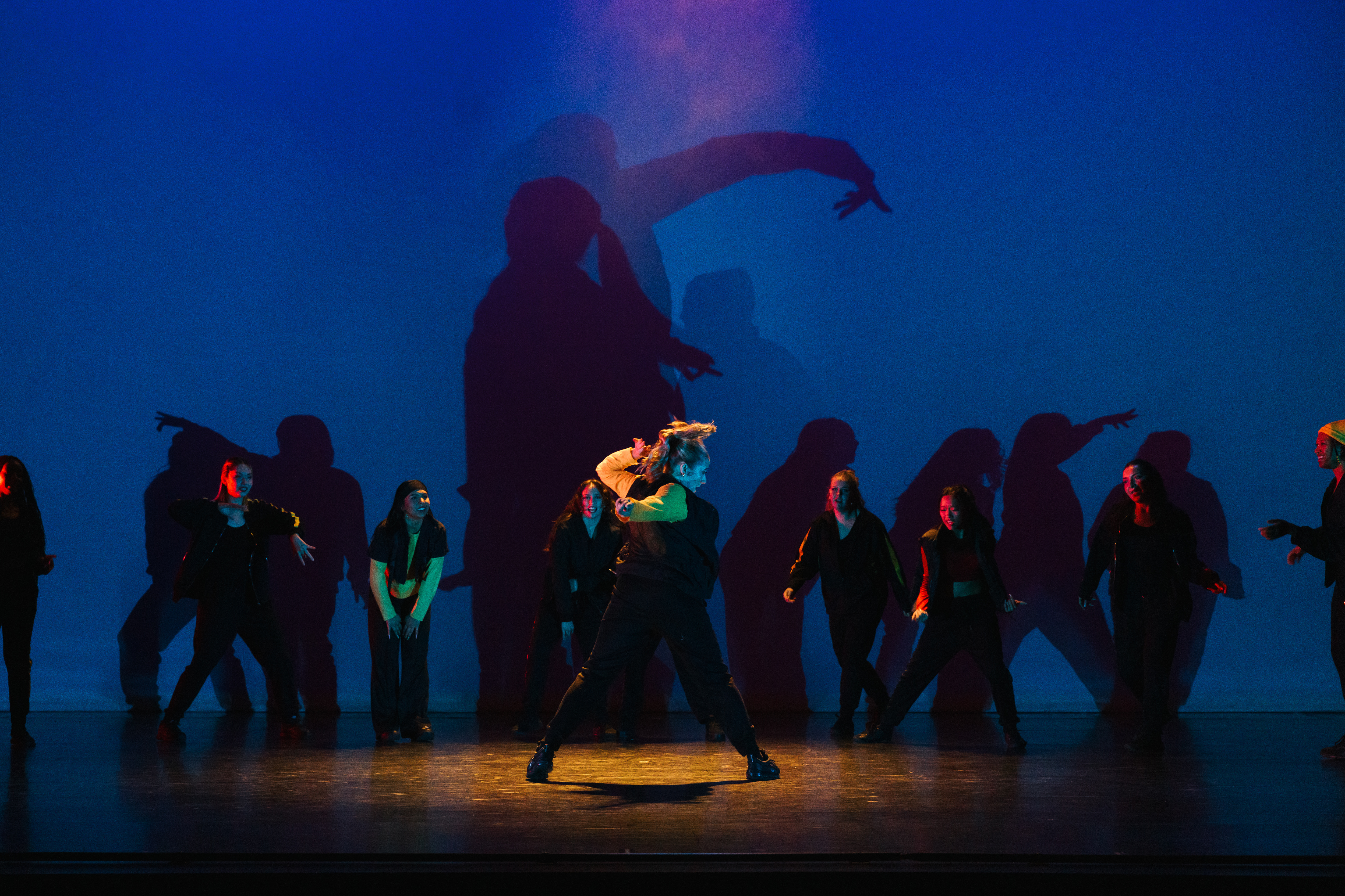 A group of dancers perform on stage under dramatic blue lighting, with bold shadows cast behind them that amplify movement and create a striking visual backdrop.