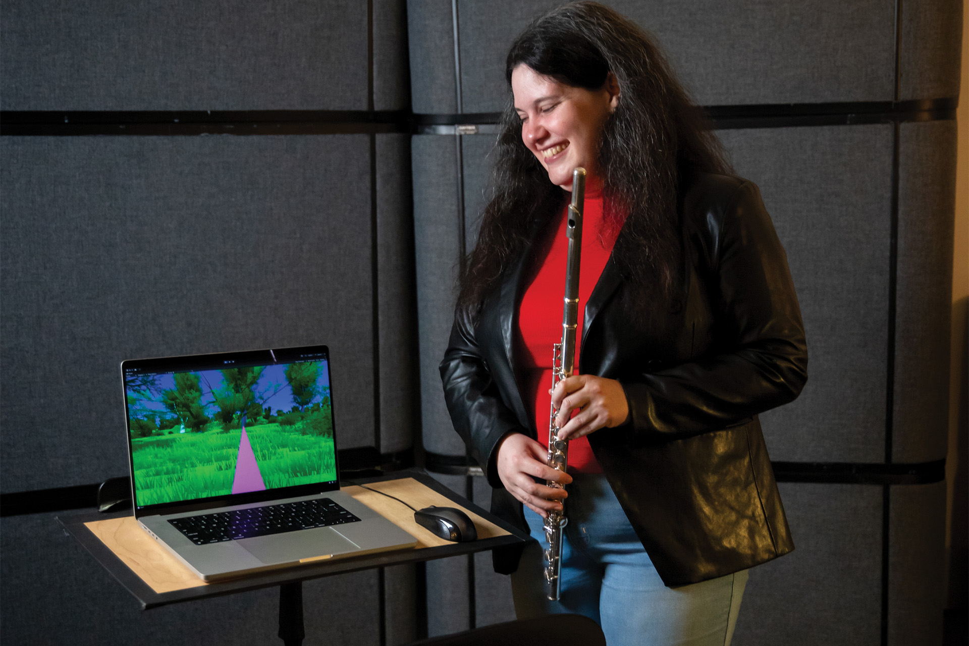 Rebecca Larkin smiles while holding a flute beside a laptop displaying a colorful virtual landscape, in a sound-treated studio space.