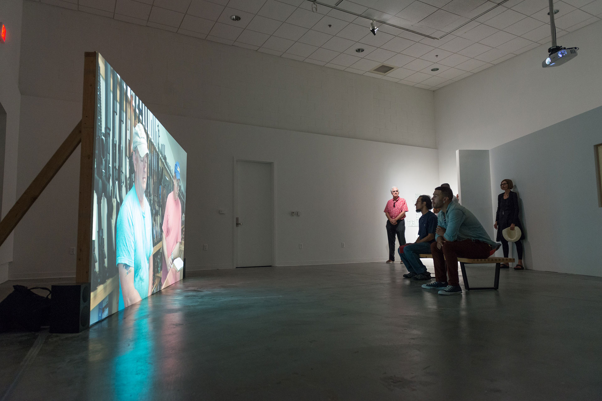 Visitors sit and stand in a gallery space watching a large wall projection, with the video casting light across the room