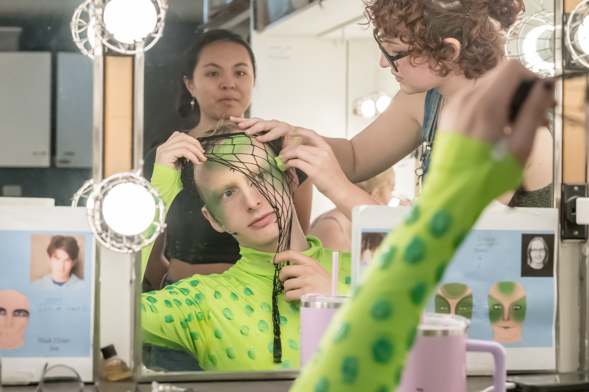 Performer in a bright green costume sits at a lighted dressing room mirror while two others assist with adjusting a netted headpiece