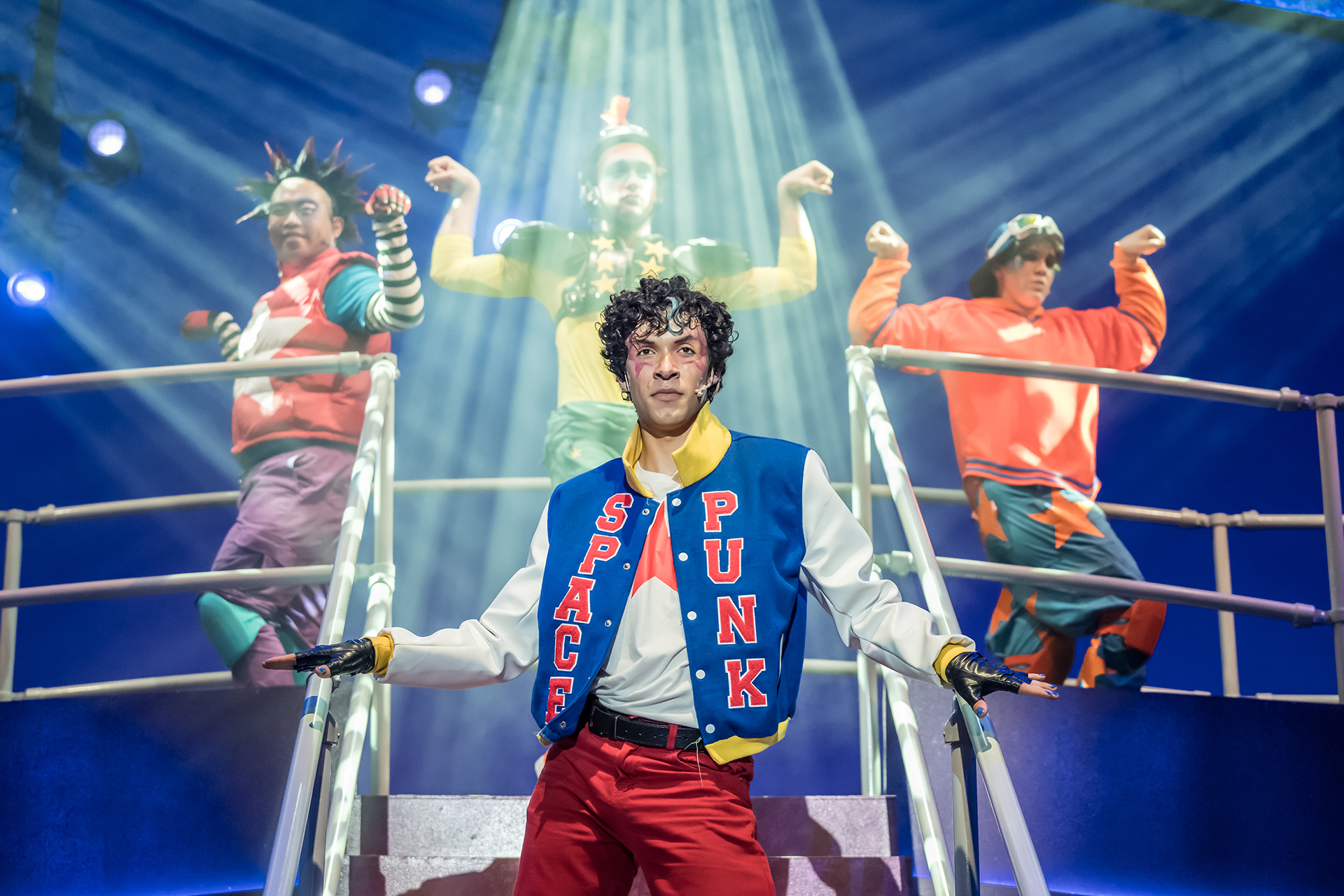 Performer stands at the front of a staged platform in a colorful costume while three performers behind pose under bright overhead stage lighting