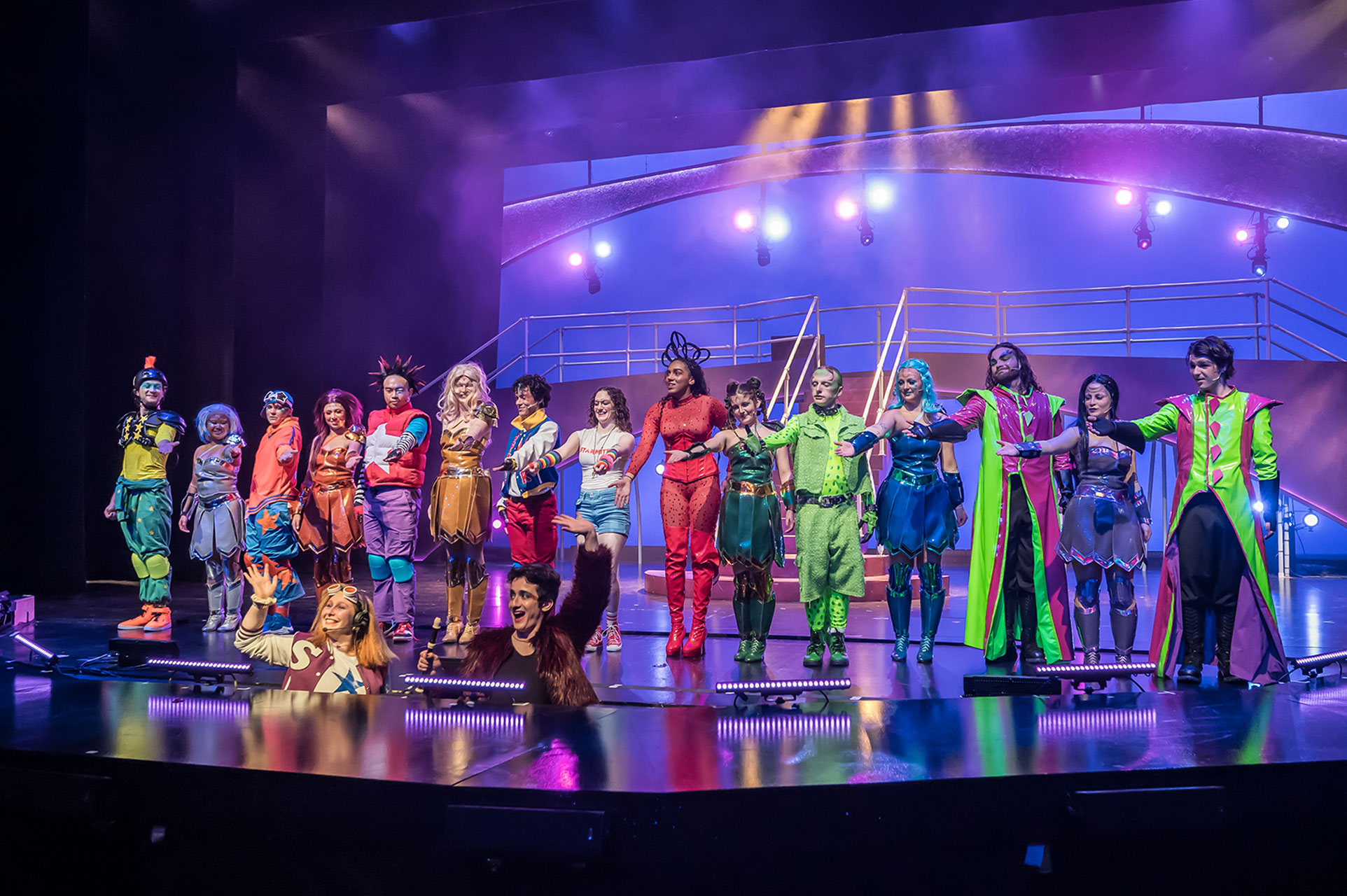 Full ensemble in bright, multicolored costumes stands in a line taking a bow on a staged platform under blue and purple theatrical lighting