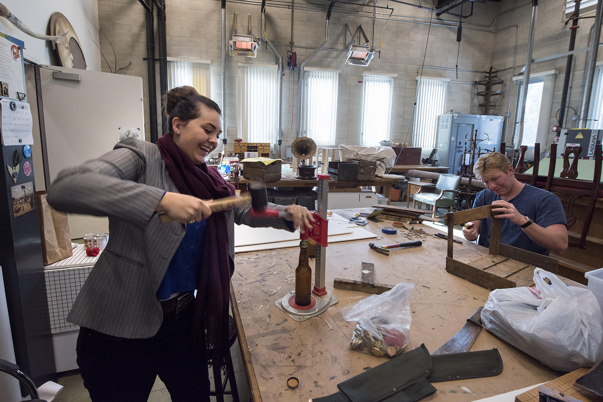 Two people work at a large table in a scenic design workshop, hammering and assembling wooden props surrounded by tools and materials