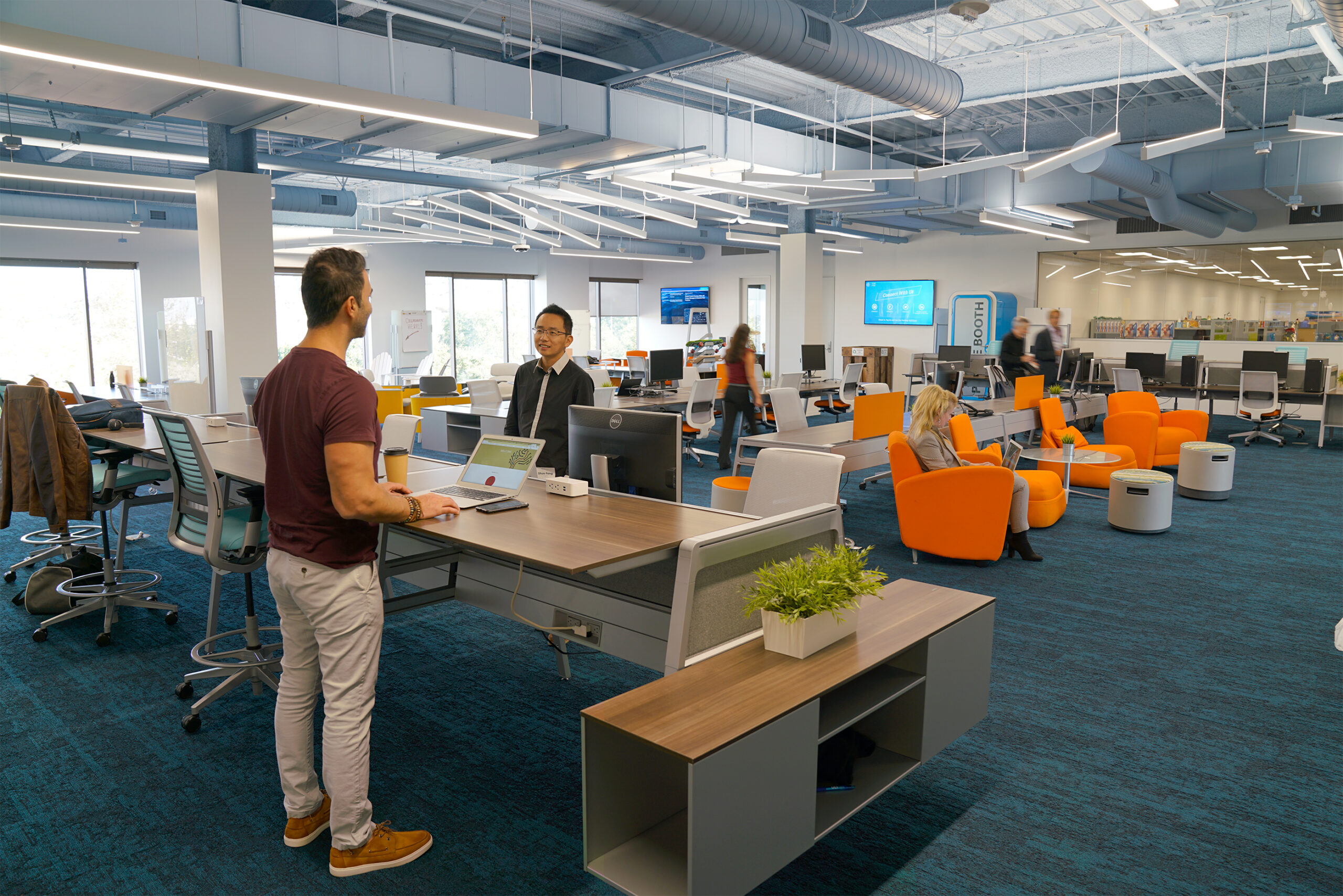 People use computers and collaborate in a modern open office with shared desks, lounge seating and bright overhead lighting