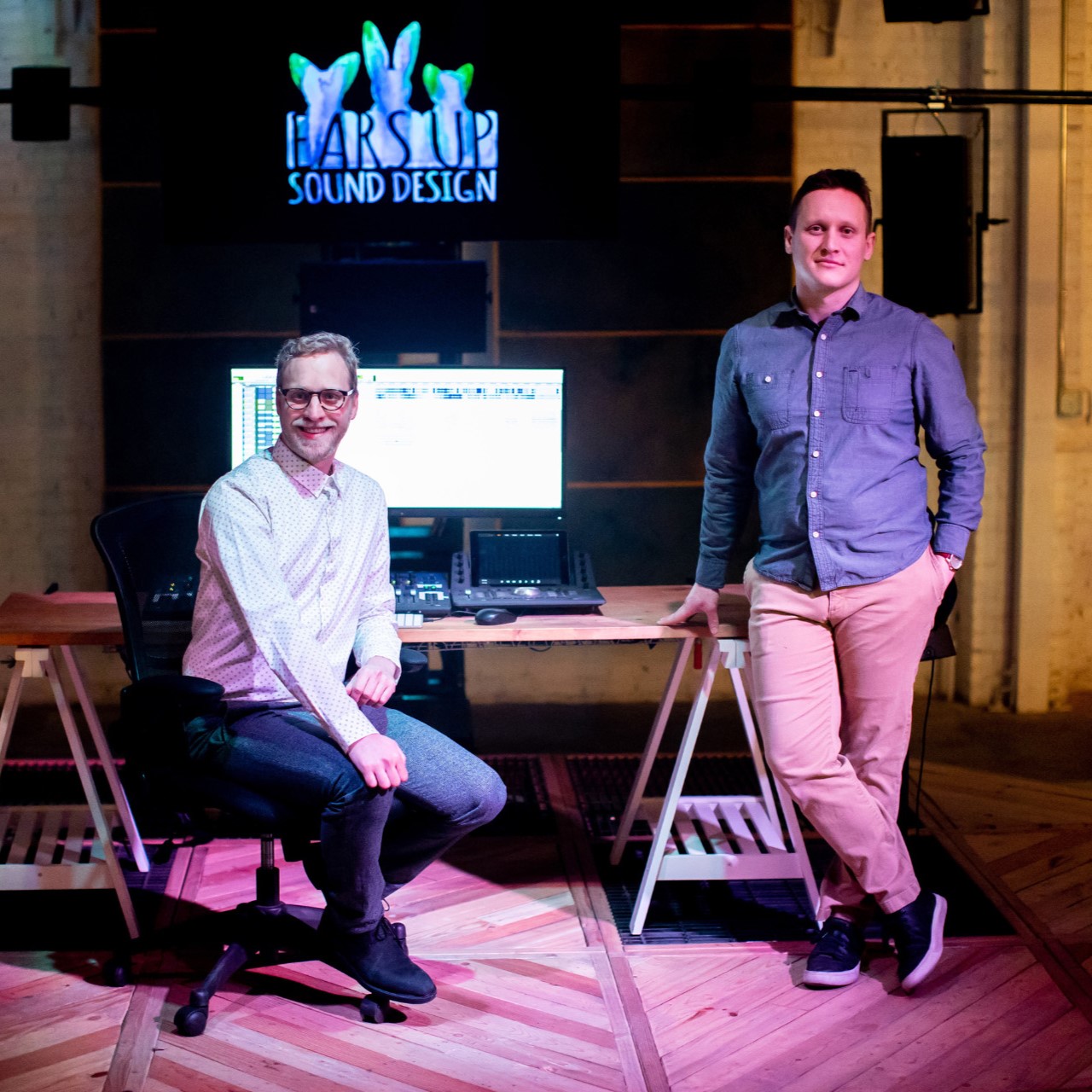 Two people pose in a sound design studio beside a computer workstation and audio equipment, with a “Farside Sound Design” sign displayed behind them