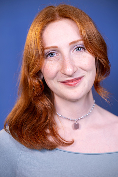 Headshot of Isabel Toteda behind a periwinkle background with red straight hair and a necklace.
