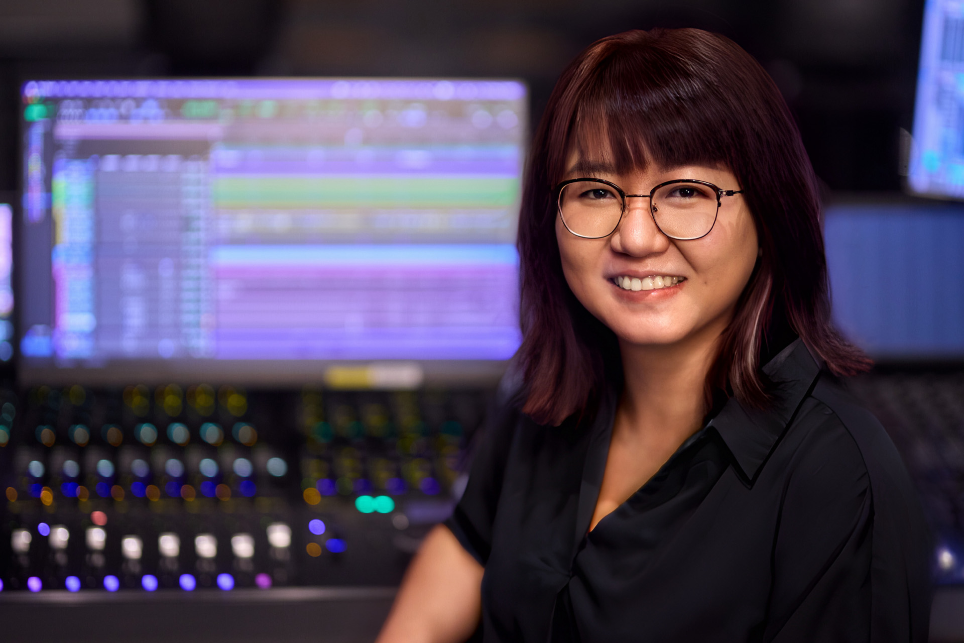Guo Ningru smiles confidently in a professional recording studio, seated in front of a mixing console with colorful audio tracks visible on a monitor screen behind her. She wears glasses and a black blouse, with her dark burgundy hair framing her face.