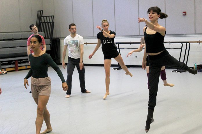 A group of dance students rehearse in a studio, performing coordinated movements with extended arms and lifted legs while focusing on technique and timing