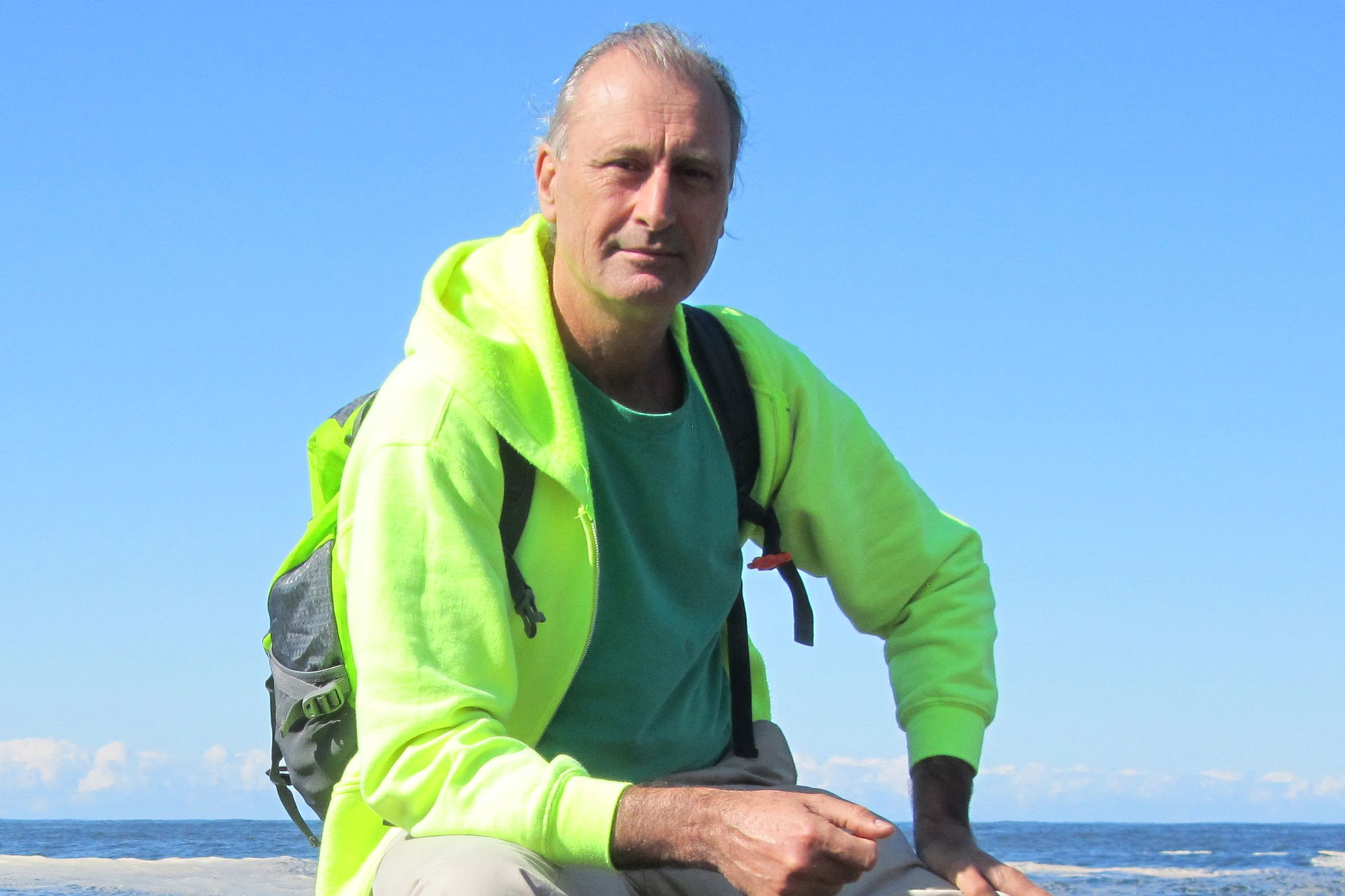 Photo of UCI Professor Simon Penny seated outdoors on a beach, wearing a neon green hoodie and backpack, with a clear blue sky and ocean behind him.