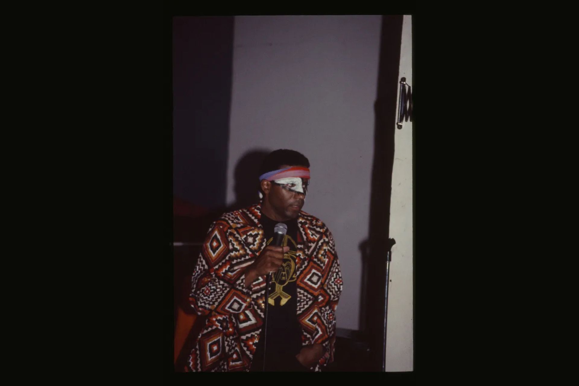 Ulysses Jenkins speaks into a microphone wearing a bold geometric-patterned jacket and a red, white, and blue headband, standing against a gray wall.