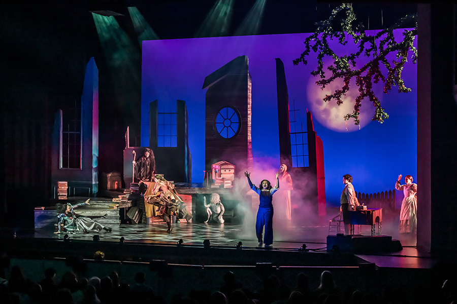 A wide shot of a theatrical stage production featuring several performers in period costumes amid atmospheric fog and dramatic purple and blue lighting. A performer in a blue outfit stands center stage with arms raised, flanked by other cast members. The set includes tall building facades, a round window and a moonlit sky backdrop with a bare tree branch overhead.