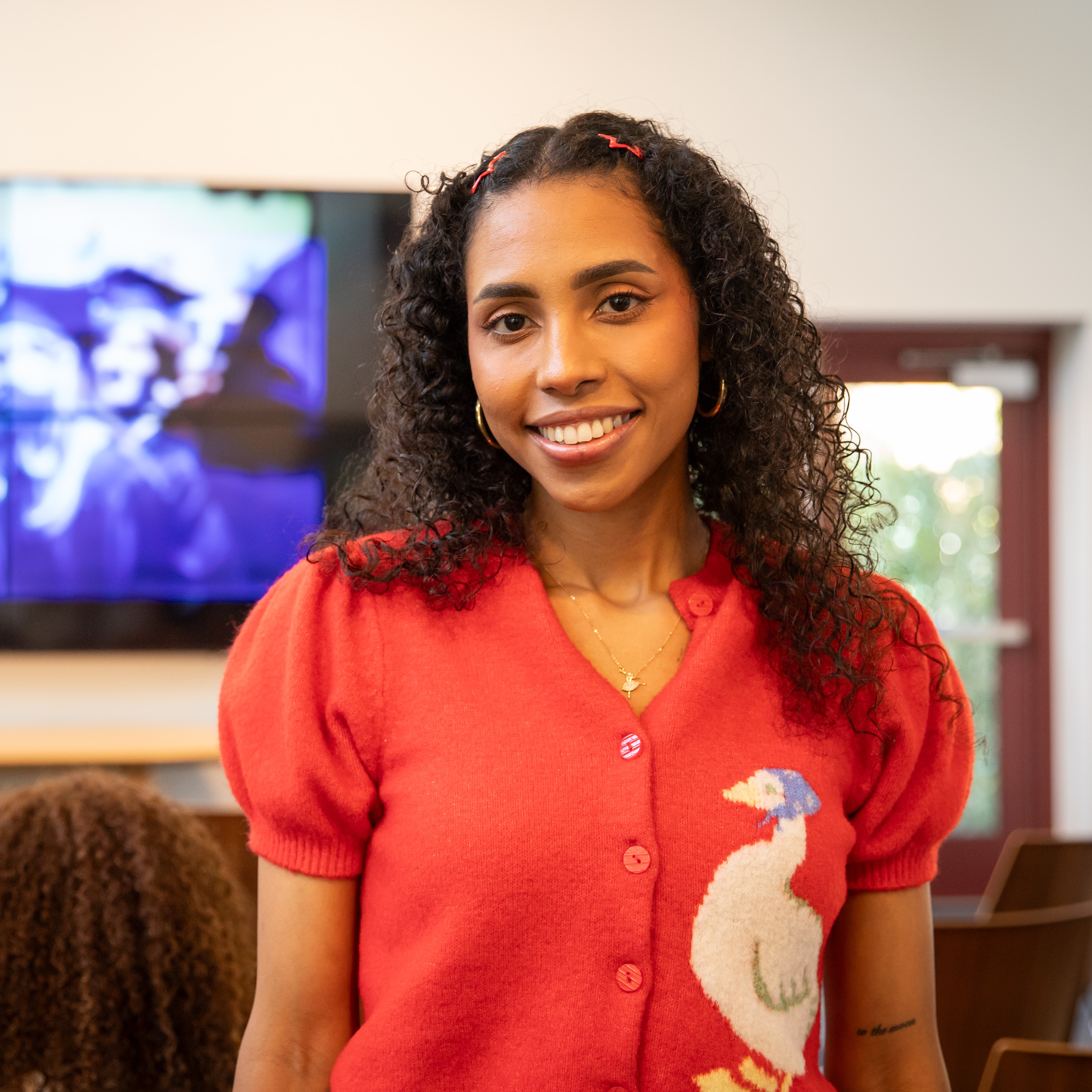 Headshot of Adanariz Baños Benitez - A smiling woman with curly hair stands in a community gathering space wearing a red cardigan with a white bird design. Digital artwork is visible on a screen behind her.