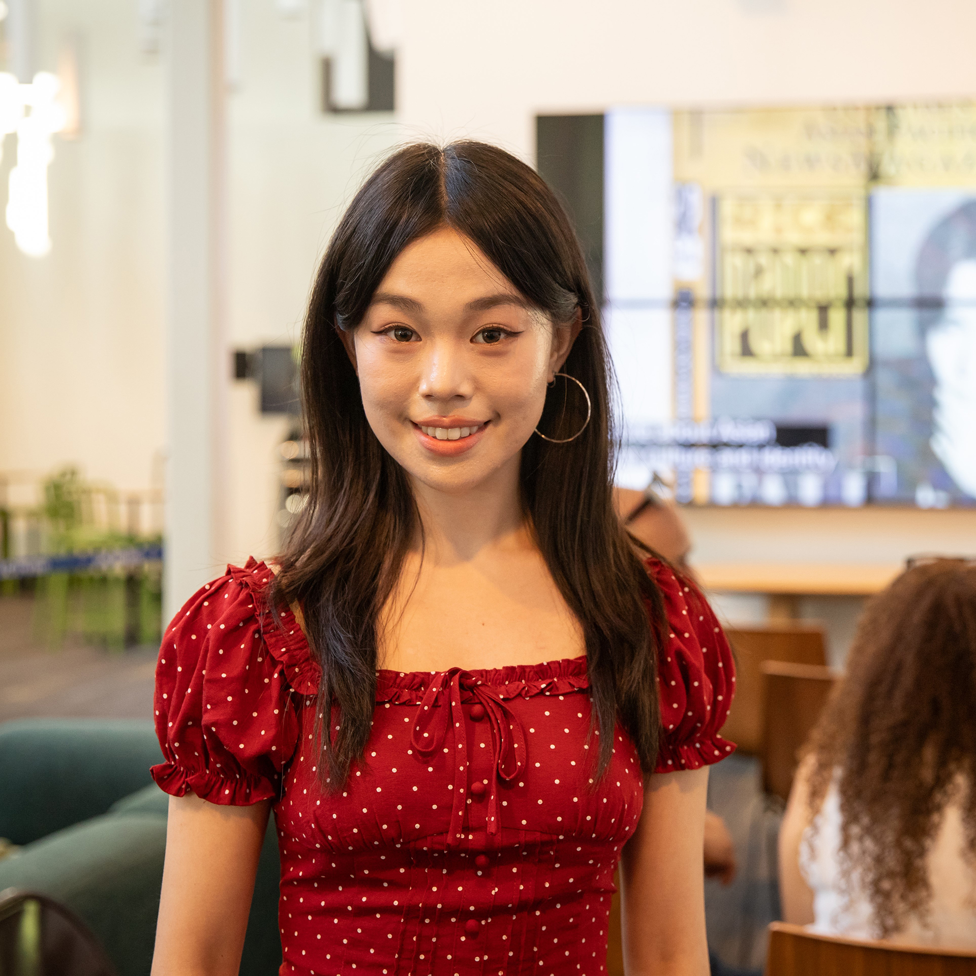 Headshot of Alina Yuan - A woman wearing a red polka-dot dress smiles toward the camera in a bright indoor gathering space. Blurred displays and attendees appear in the background.
