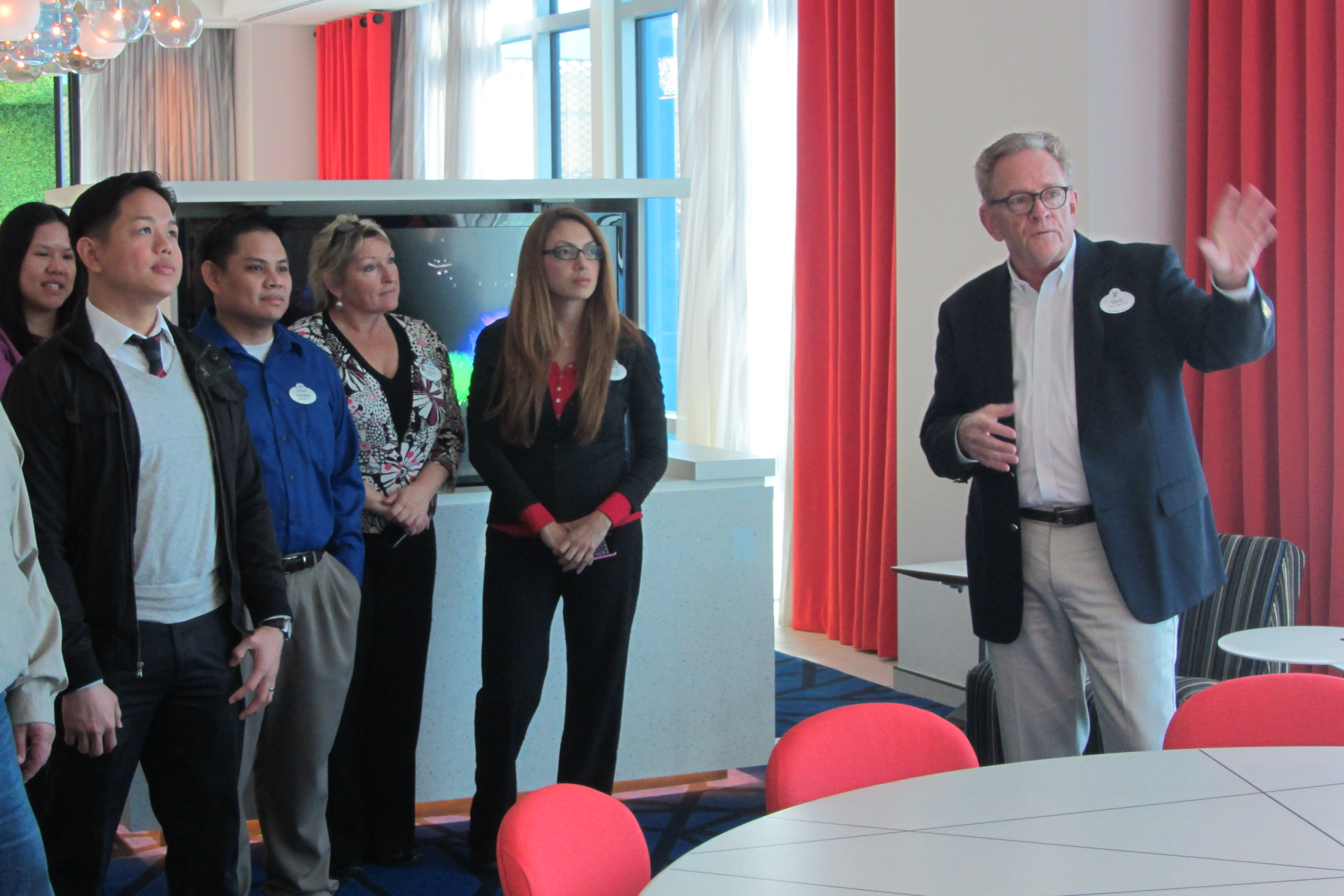 David Golbeck in his role at Disney Imagineering (A group of attendees listens as a speaker gestures during a guided presentation in a modern conference room with floor-to-ceiling windows and red accent curtains)