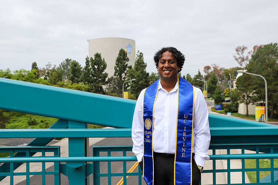 Hitesh Benny, a UC Irvine Class of 2026 graduate, smiles while standing on a teal pedestrian bridge, wearing a white button-up shirt and a blue and gold UC Irvine graduation stole, with trees and a campus water tower behind him.