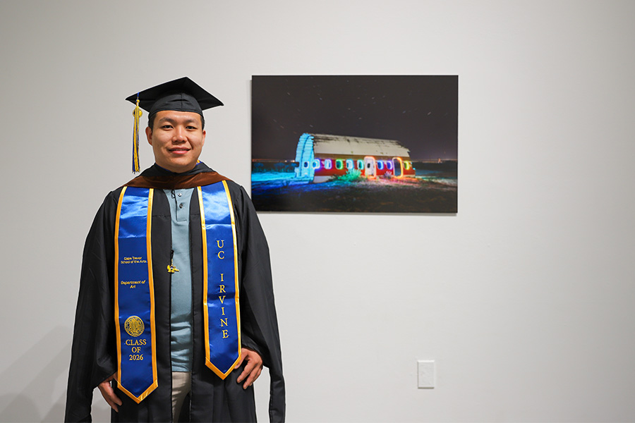 Yuzhe Zhang, a UC Irvine Class of 2026 graduate, stands in his black cap and gown and blue and gold UC Irvine stole beside one of his photographs, which shows a colorfully lit structure against a dark night sky in a gallery setting.