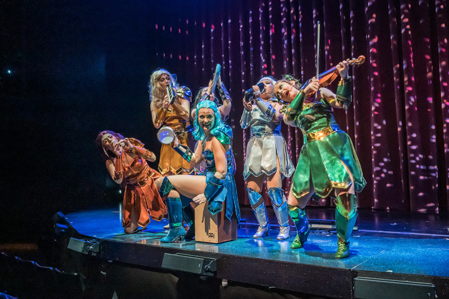 Five performers in colorful, metallic warrior-style costumes — in orange, blue, silver, and green — strike dynamic poses on a dimly lit stage during a production of 'Starmites' at the Claire Trevor School of the Arts. One performer kneels at center playing a cajón while others play a violin, horn, and other instruments under purple and pink stage lighting.