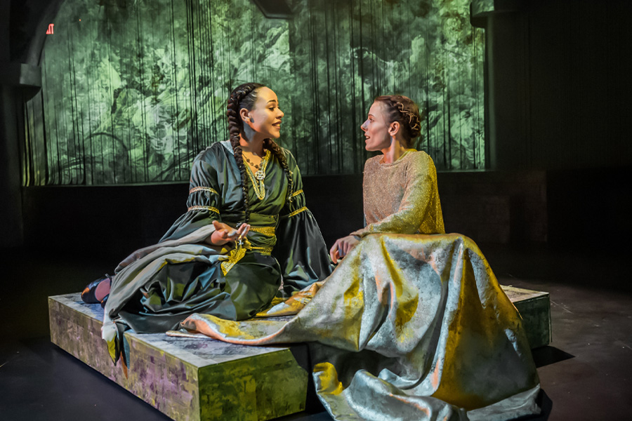 Two performers in medieval-style gowns sit facing each other on a low platform during a production of The Green Knight at the Claire Trevor School of the Arts. One wears a dark green dress with gold trim and a long braid; the other wears a shimmering gold and silver gown. They are lit against a backdrop of projected forest imagery in green tones."